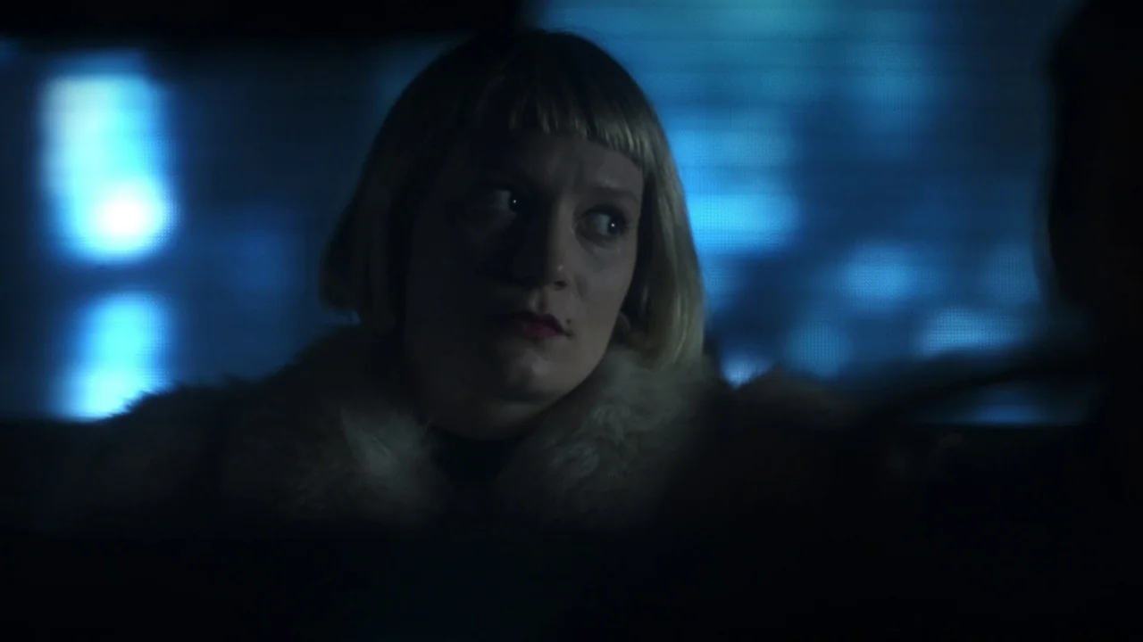 Piercing (2018) movie still: closeup — A dark interior of a vehicle at night featuring a bright blue screen reflection. A blonde woman with blunt…