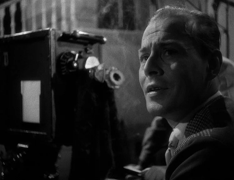Prison (1949) movie still: closeup — A man sits next to a vintage professional film camera in a dark space. The man looks intently towards the right…
