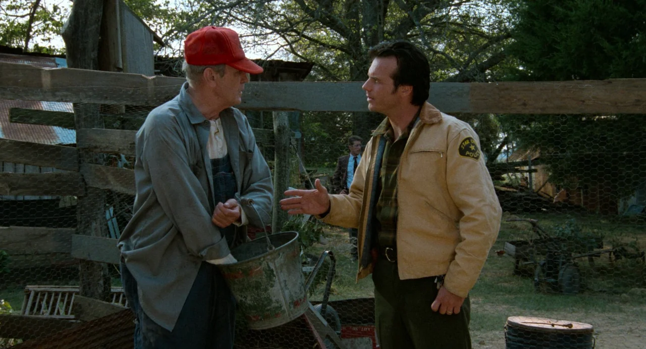 One False Move (1991) movie still: medium — A farmyard setting featuring a wooden wire fence and scattered rustic equipment. Two men stand in the…