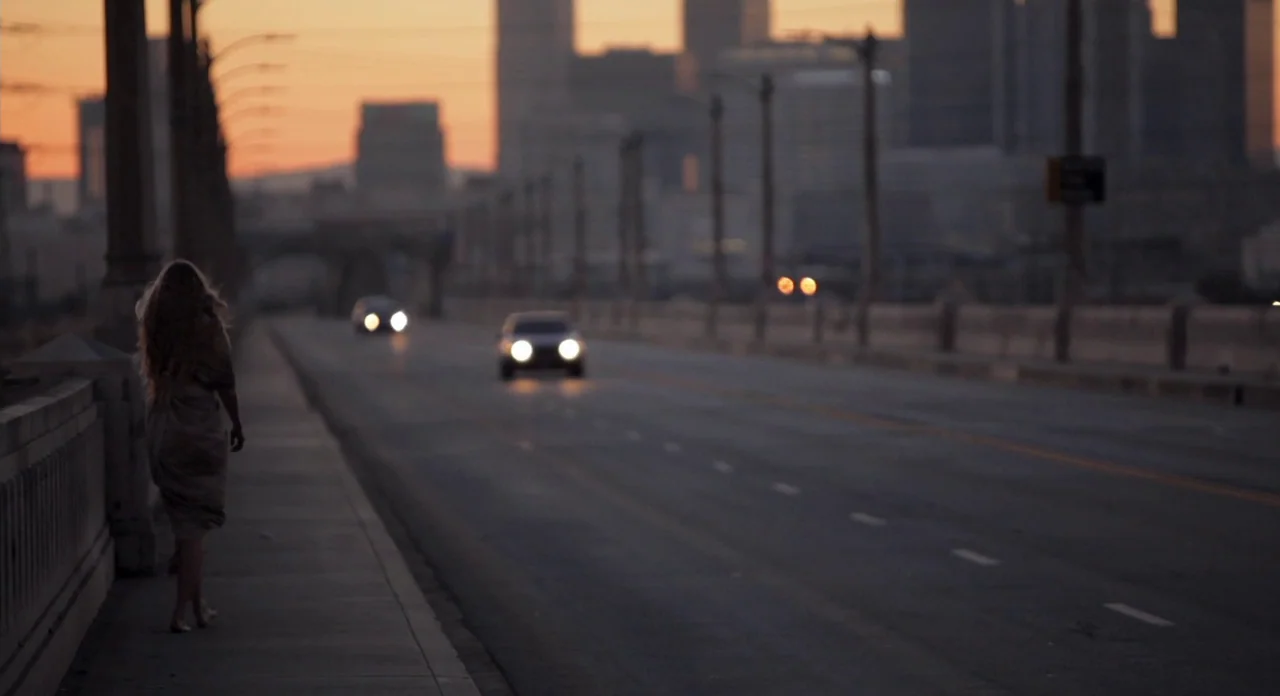 Sound Of My Voice (2011) movie still: fullbody — A wide concrete bridge spans across a hazy urban skyline during sunset. A solitary woman with long…