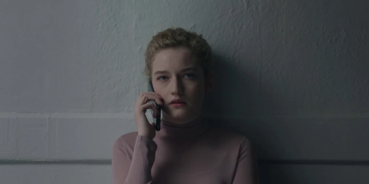The Assistant (2019) movie still: medium — A woman leans against a textured gray concrete wall in a dimly lit space. She holds a thin black…