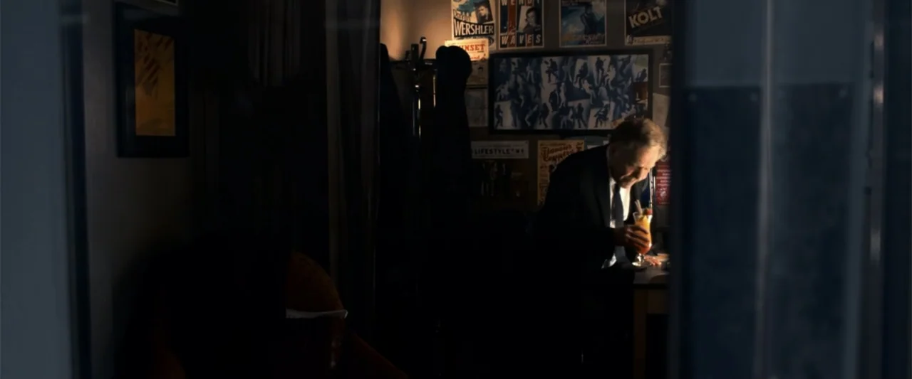 2012 (2009) movie still: medium — A dark office space is filled with framed posters and documents on the wall. An older man sits alone at his desk…