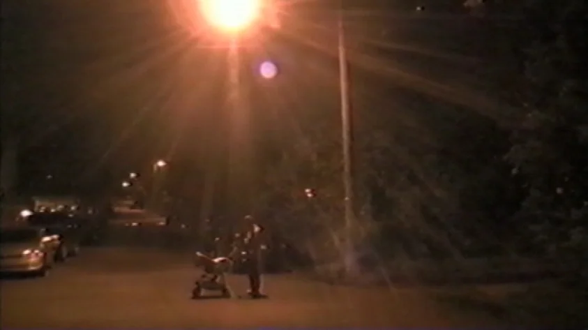 Trash Humpers (2009) movie still: wide — A dark city street is illuminated by a single glowing overhead streetlamp at night. A person stands on the…