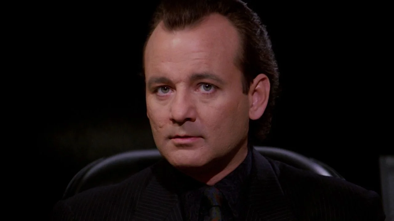 Scrooged (1988) movie still: closeup — A dark, featureless studio setting with a subject centered in the frame. The man sits in a chair, facing the…