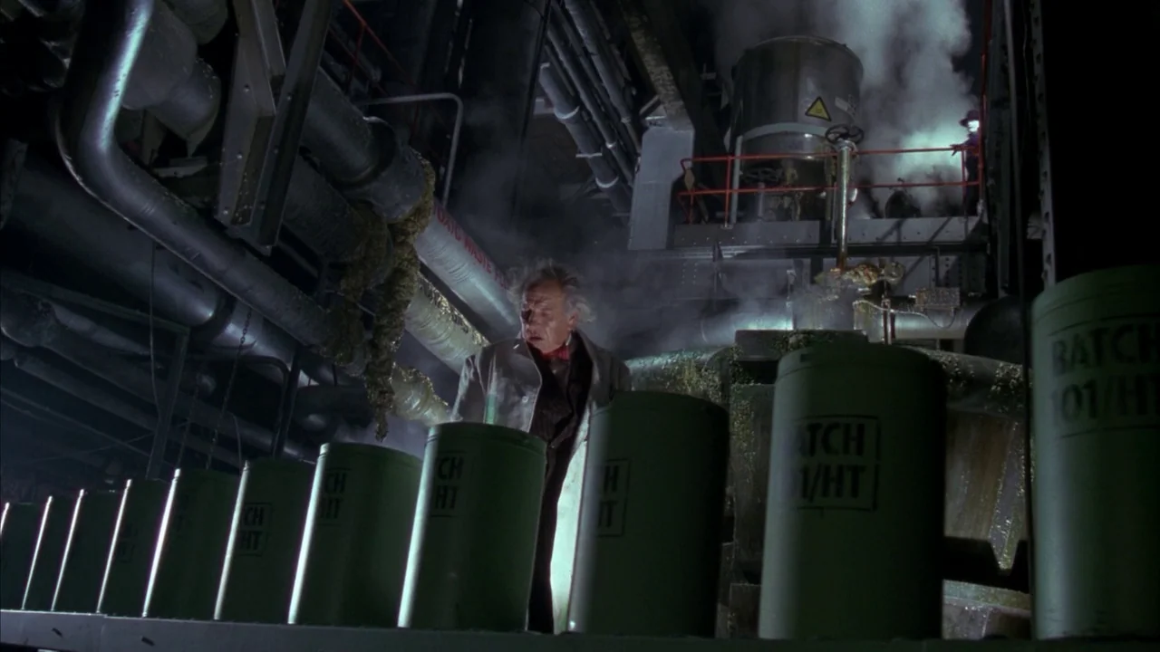 Batman (1989) movie still: medium — An industrial facility features rows of green barrels and complex overhead piping and machinery. An older man in…