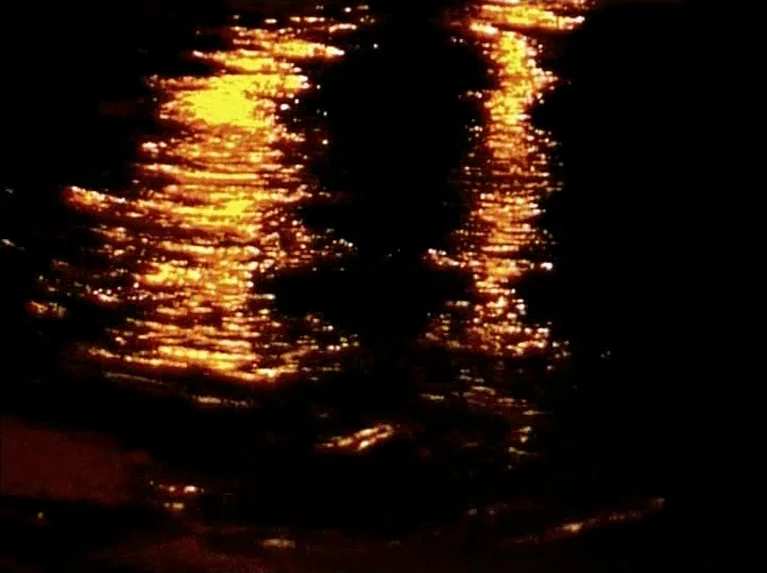 Eloge de L’amour (2001) movie still: establishing — The surface of a dark body of water at night captures bright yellow light. Ripples break the…