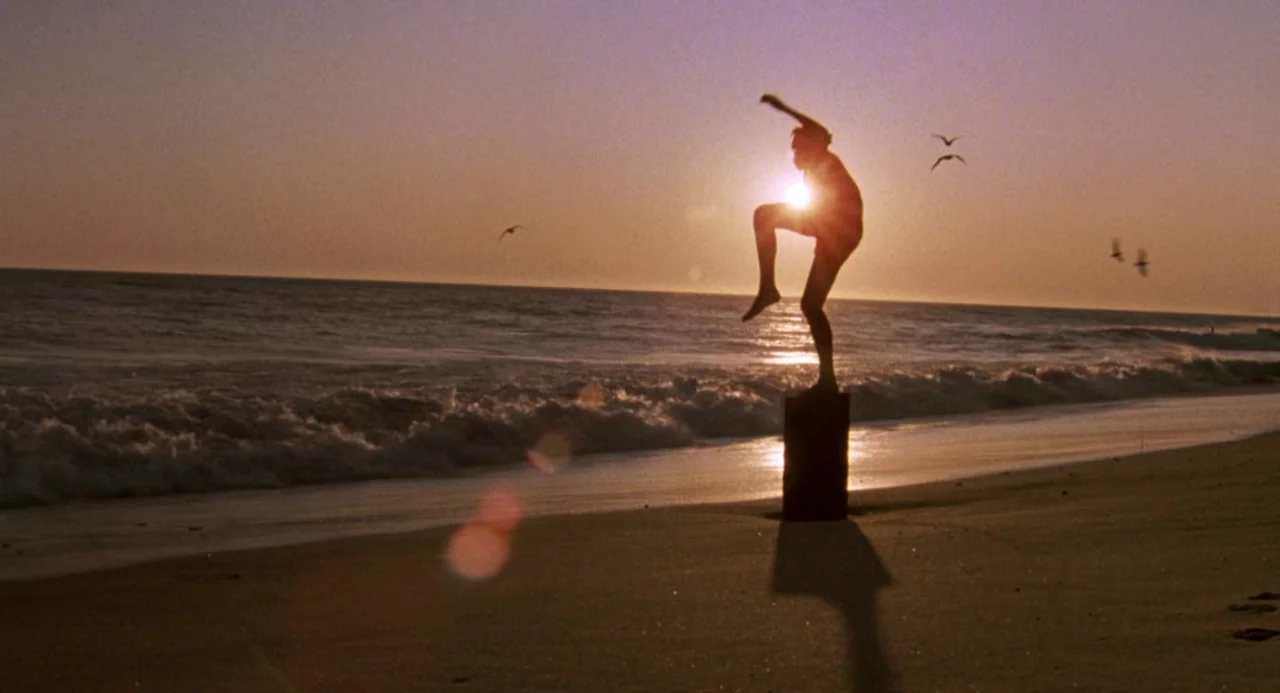 The Karate Kid (1984) movie still: wide — A sandy shore during sunset features gentle waves and a single wooden post. A man stands on the post in a…