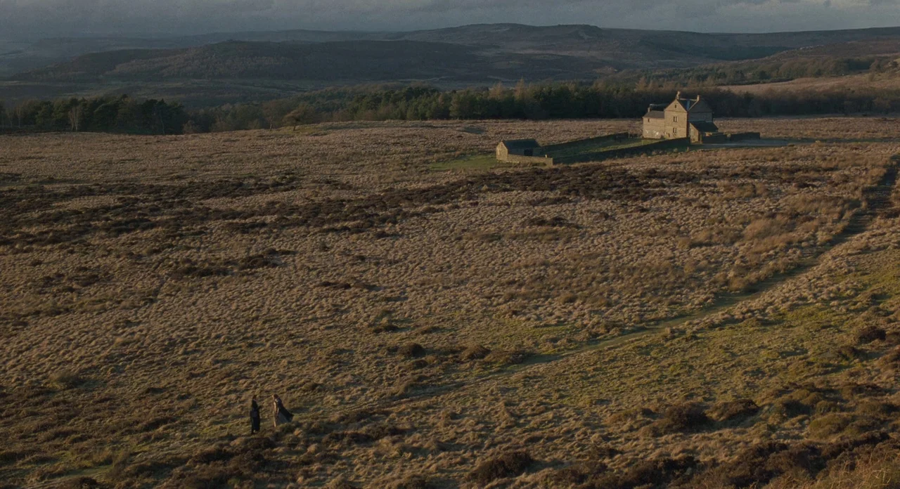 Jane Eyre (2011) movie still: wide — A vast, expansive moorland landscape stretches under an overcast sky toward a stone building. Two figures stand…