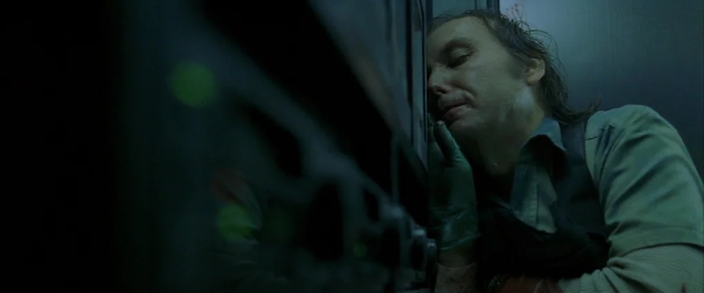 Panic Room (2002) movie still: medium — A dimly lit industrial environment featuring a metallic fuel pump in the foreground. A man in work clothes…