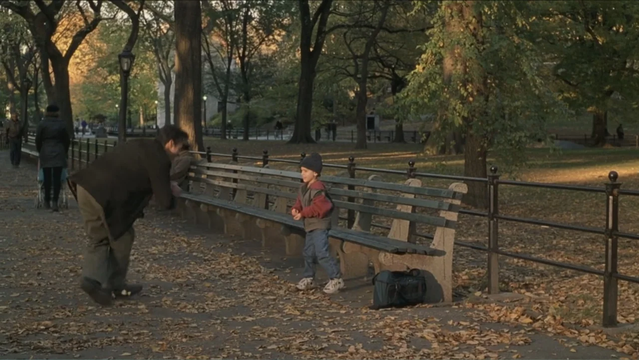 Big Daddy (1999) movie still: fullbody — An autumnal city park features large trees, fallen leaves, and wooden benches with iron frames. A man in a…