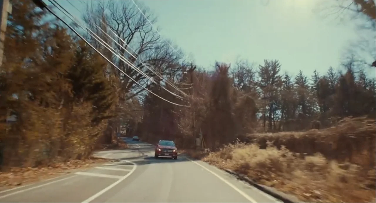 An Imperfect Murder (2017) movie still: wide — A narrow paved road winds through a dense area of bare autumn trees. A small red vehicle travels…