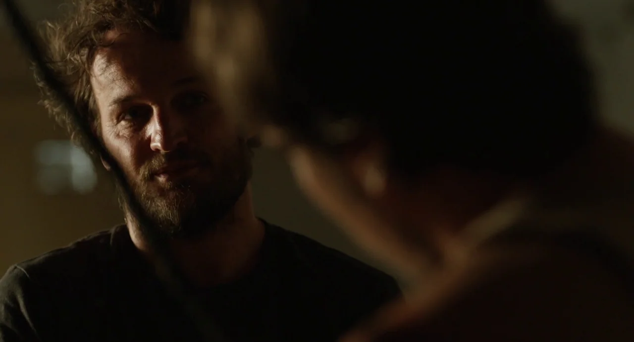 Zero Dark Thirty (2012) movie still: closeup — A dimly lit room with an out-of-focus individual in the foreground. A man with curly hair and a beard…