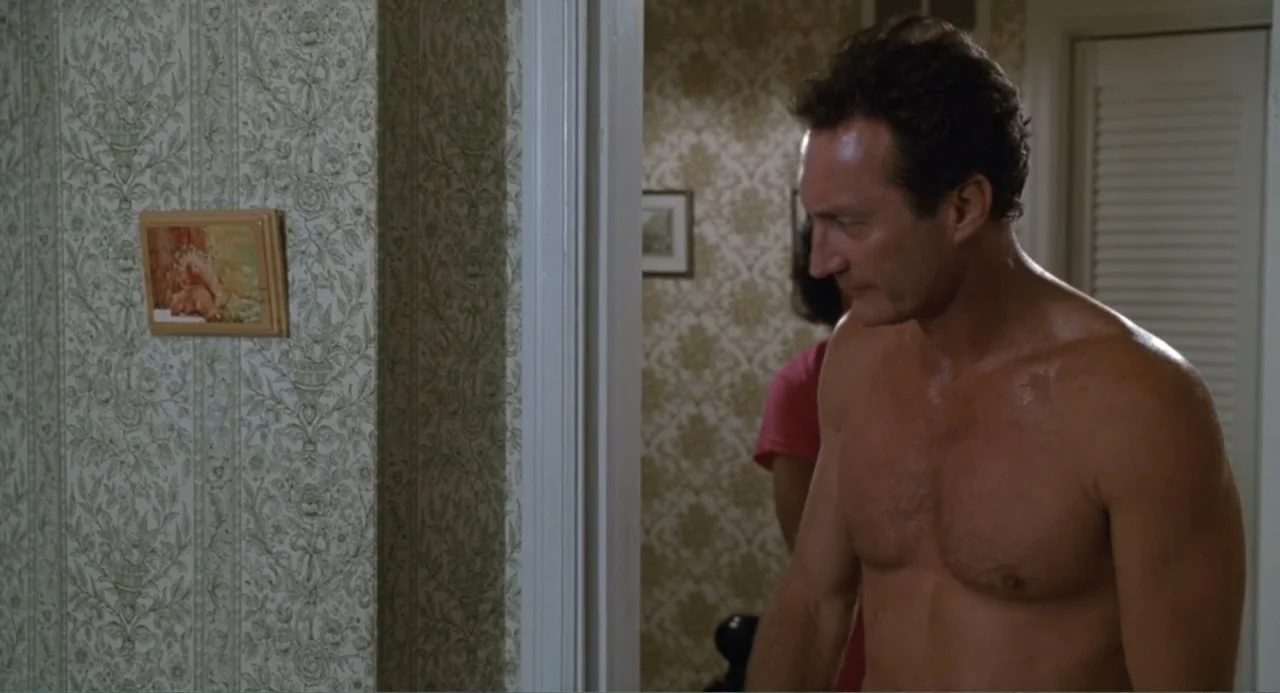 F/X2 (1991) movie still: medium — A residential interior features wallpaper with a floral damask pattern and a framed picture. A shirtless man…