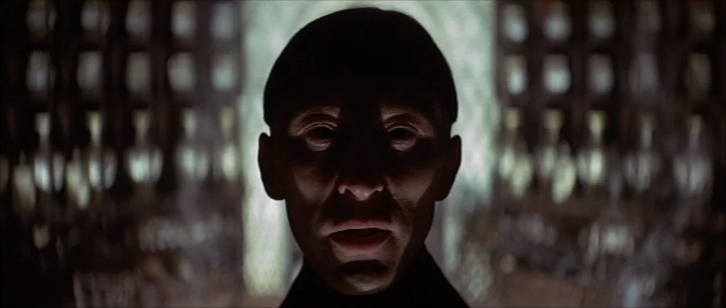 The Devils (1971) movie still: closeup — An interior space features tall, blurred rows of vertical objects in the background. A man stands centered…