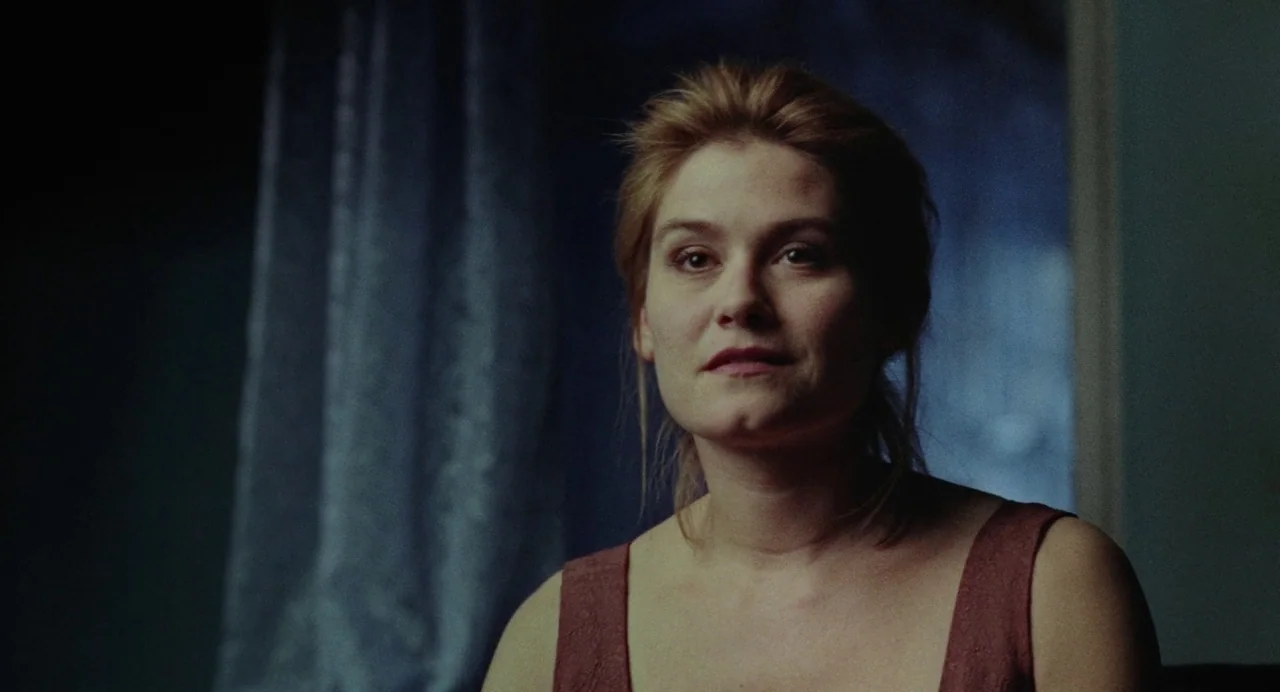 What Happened Was… (1994) movie still: medium — A dimly lit room with a dark blue wall and a draped curtain behind. A woman sits facing the camera…
