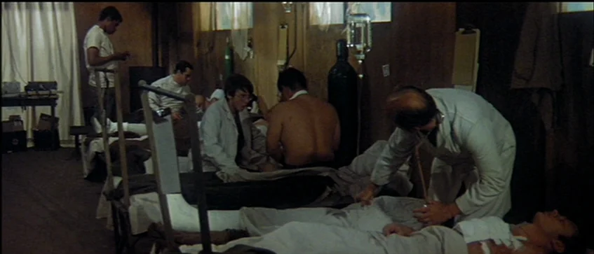 MASH (1970) movie still: wide — A crowded hospital ward features multiple beds arranged in rows with wood paneled walls. Medical staff in white…