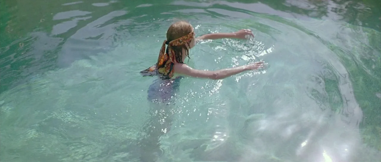 Dogtooth movie still: medium — A woman swims in a clear, bright turquoise pool with visible rippling water surfaces. The woman moves away from the…