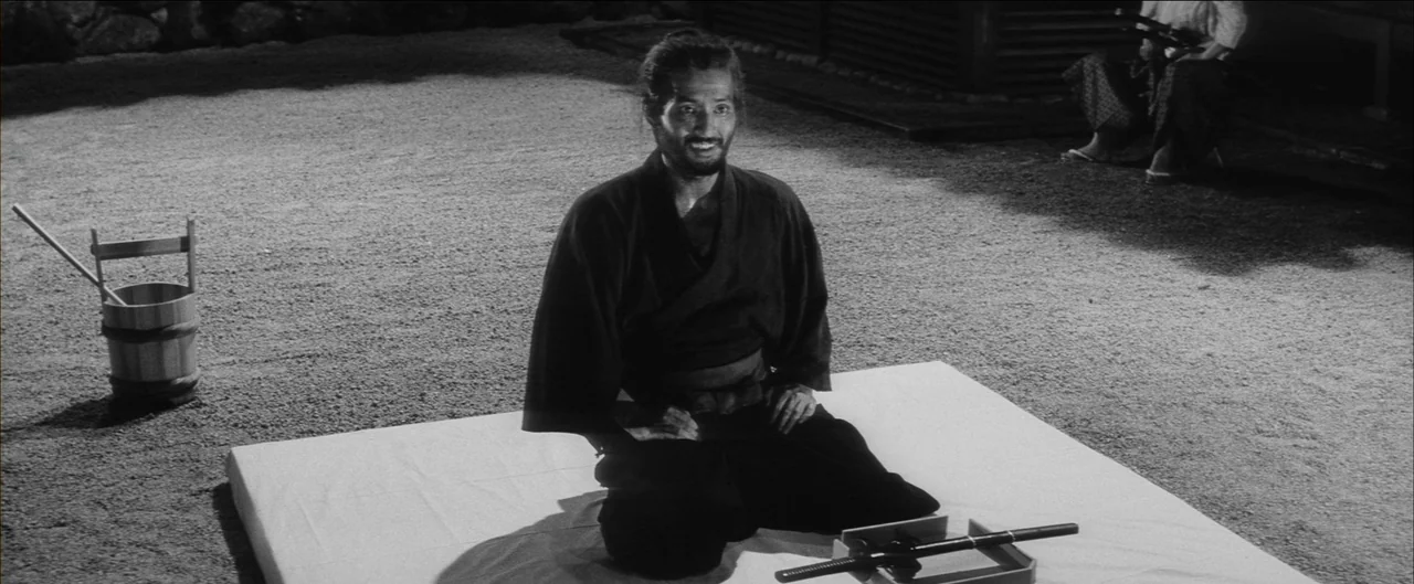 Harakiri (1962) movie still: medium — A gravel-covered courtyard at night featuring a man sitting on a white mat. A wooden bucket stands to his left…