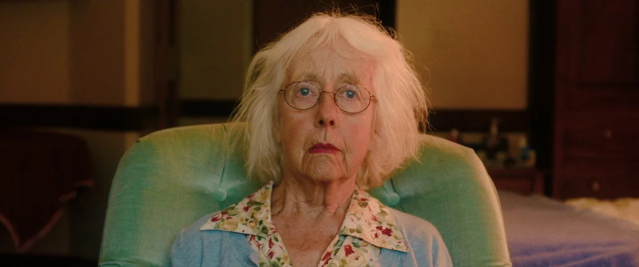 I Care A Lot (2020) movie still: closeup — An elderly woman sits in a pale green upholstered chair inside a room. She looks directly at the camera…