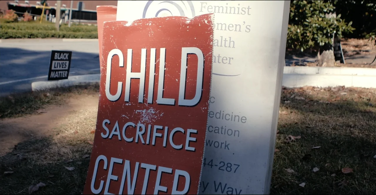 The Devil Is Busy (2024) movie still: closeup — An outdoor sidewalk area with a red sign and an adjacent health center sign. A red sign with white…