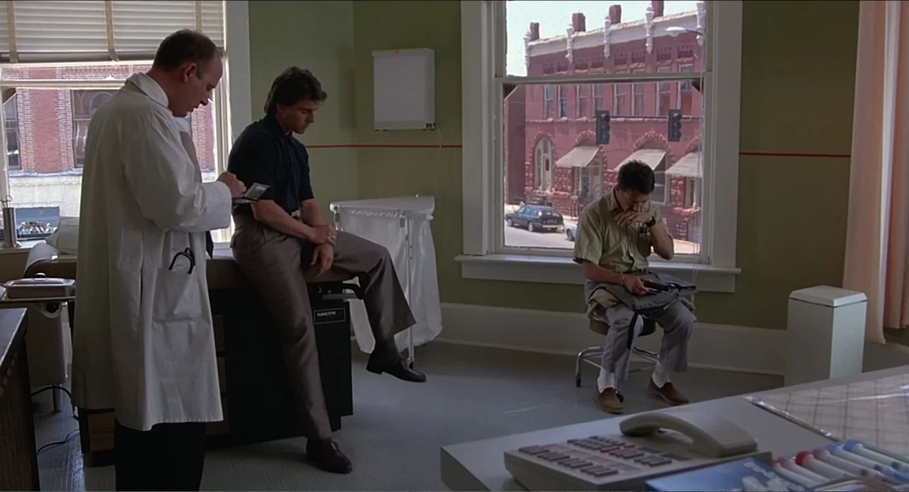 Rain Man (1988) movie still: medium — A medical exam room features pale green walls and a large window facing town. A doctor stands with a clipboard…