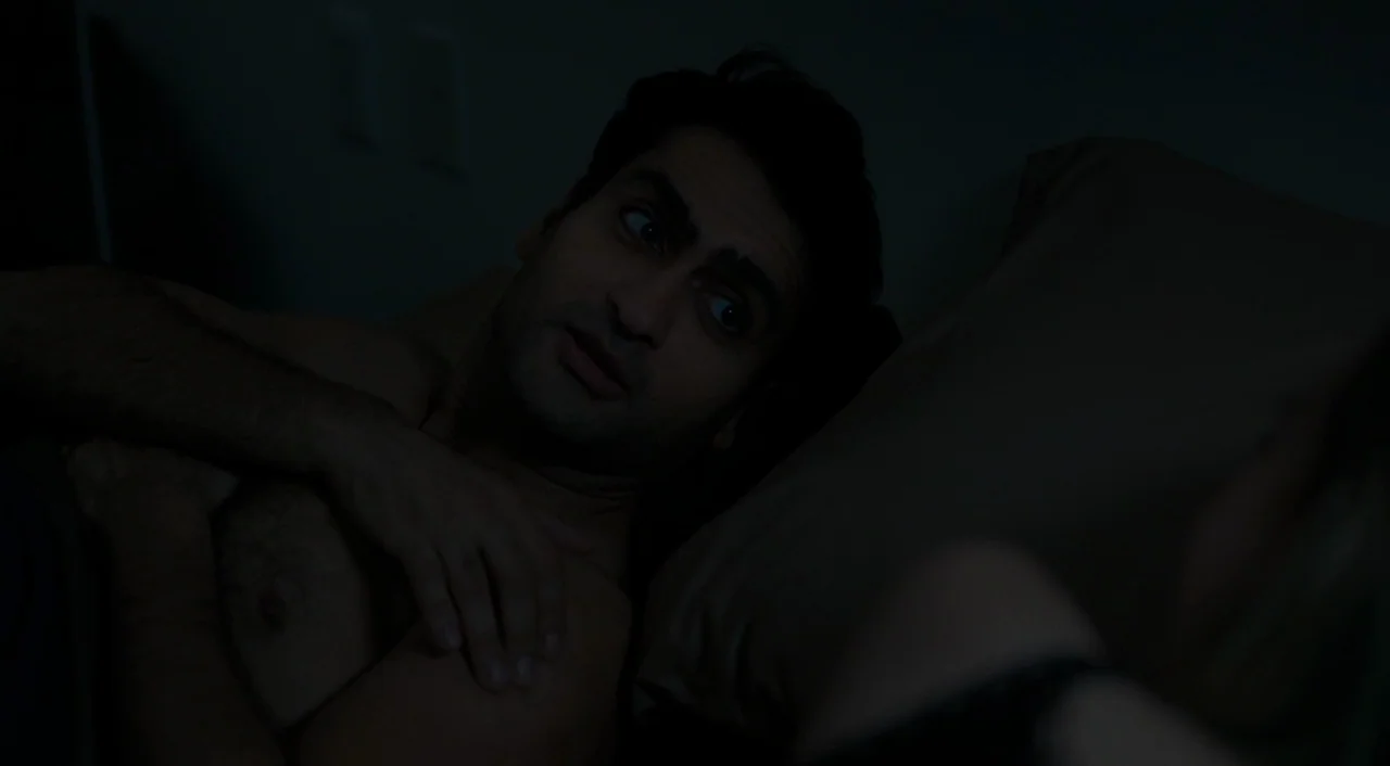 The Big Sick (2017) movie still: closeup — A man lies in a dark, dimly lit room with bedding visible beside him. The man is awake, glancing toward…