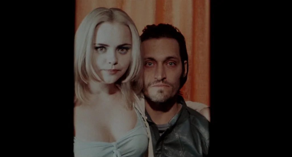 Buffalo ’66 (1998) movie still: closeup — A confined space features a textured, vertically pleated orange background curtain. A woman and a man…