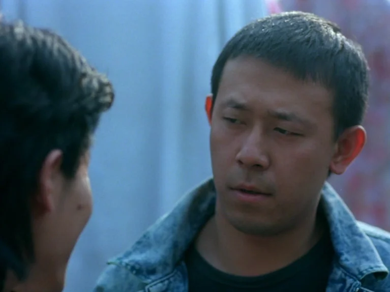 Black Snow (1990) movie still: closeup — An outdoor city setting serves as the backdrop for a conversation between two people. One man wearing a…
