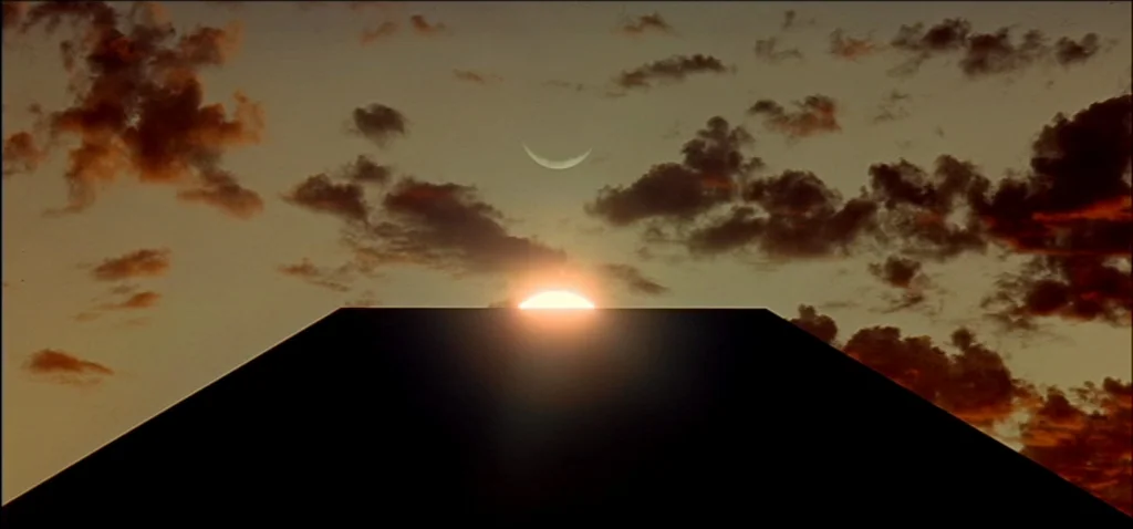 2001: A Space Odyssey (1968) movie still: establishing — A dark, rectangular monolith stands against a horizon filled with clouds at sunrise. The…
