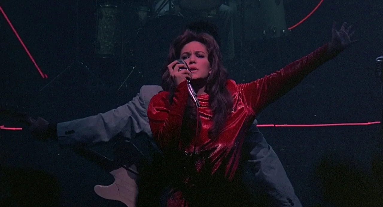 Streets of Fire (1984) movie still: medium — A dark concert venue is illuminated by thin, horizontal red neon light bars. A woman stands centrally…