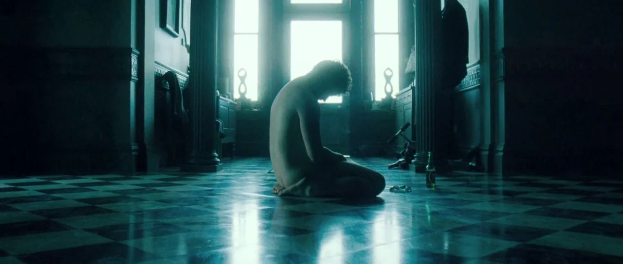 Filth (2013) movie still: fullbody — An interior hallway features a high-contrast black and white checkered tile floor. A nude man kneels on the…