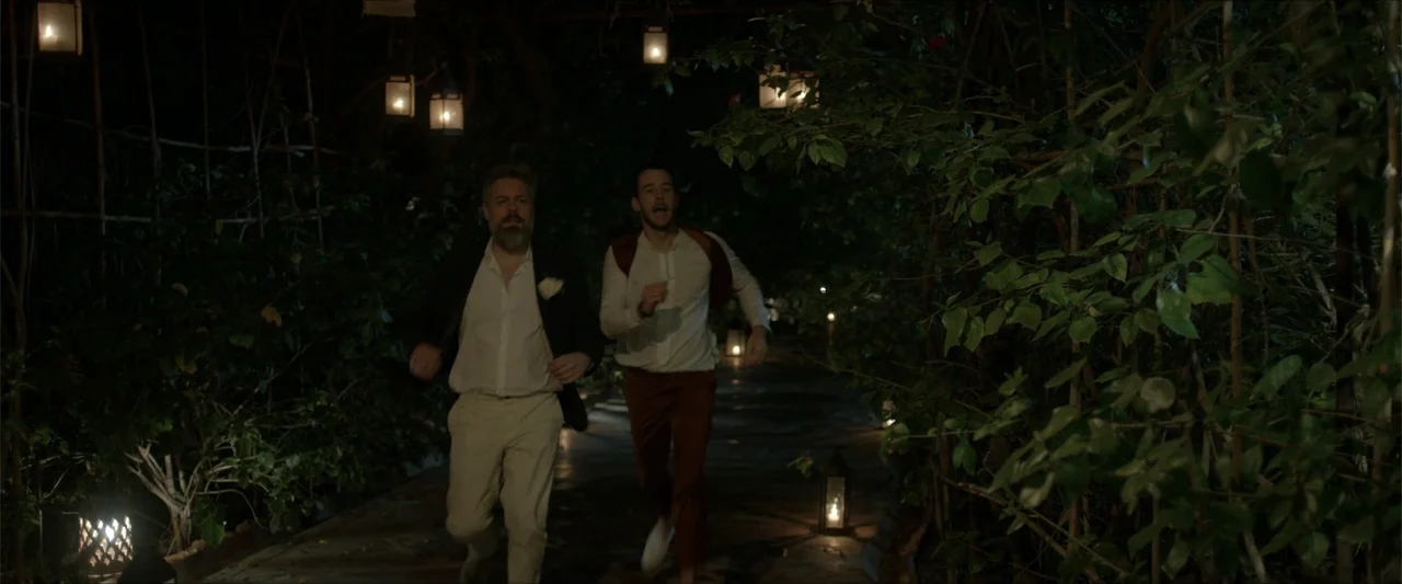 An Endless Wedding (2025) movie still: fullbody — A dark outdoor path is lined with lush foliage and hanging glass lanterns. Two men run toward the…