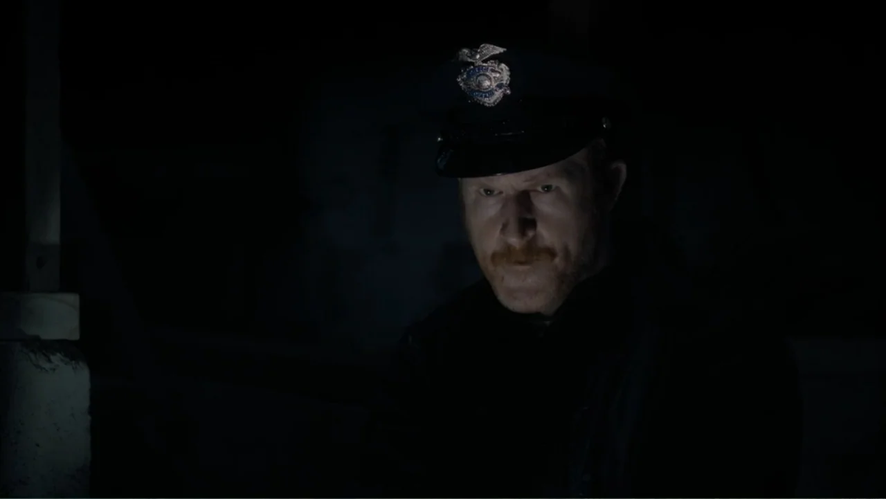 Storm Damage (2000) movie still: closeup — A dimly lit interior space with an indistinct dark background. A man in a police uniform gazes ahead…