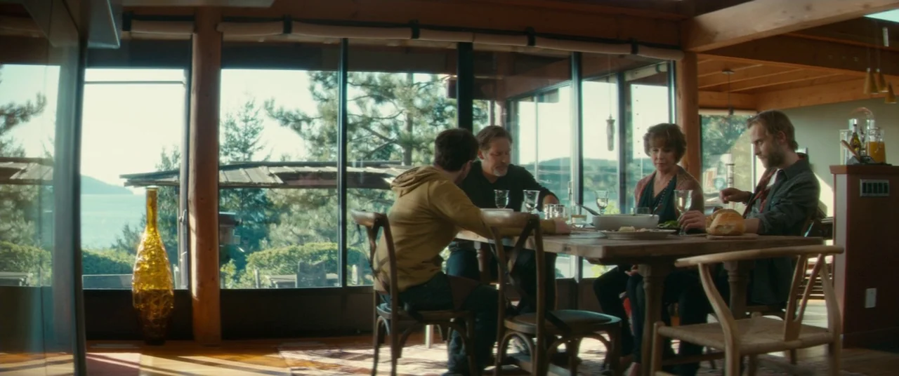 Horns (2013) movie still: wide — A modern dining room with wood framing and floor-to-ceiling windows overlooking a water view. Four individuals sit…