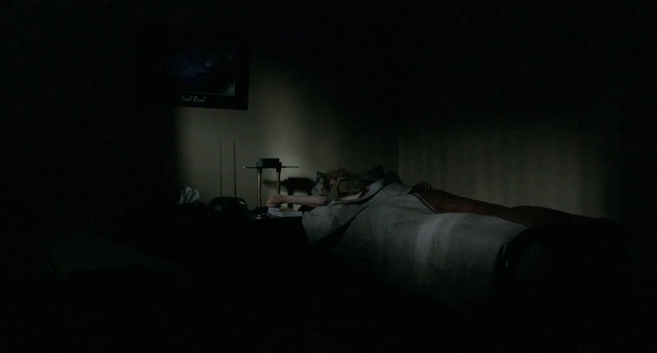 Tom at the Farm (2013) movie still: wide — A dimly lit room contains a bed with a person under a neutral-colored blanket. A bedside lamp casts a…