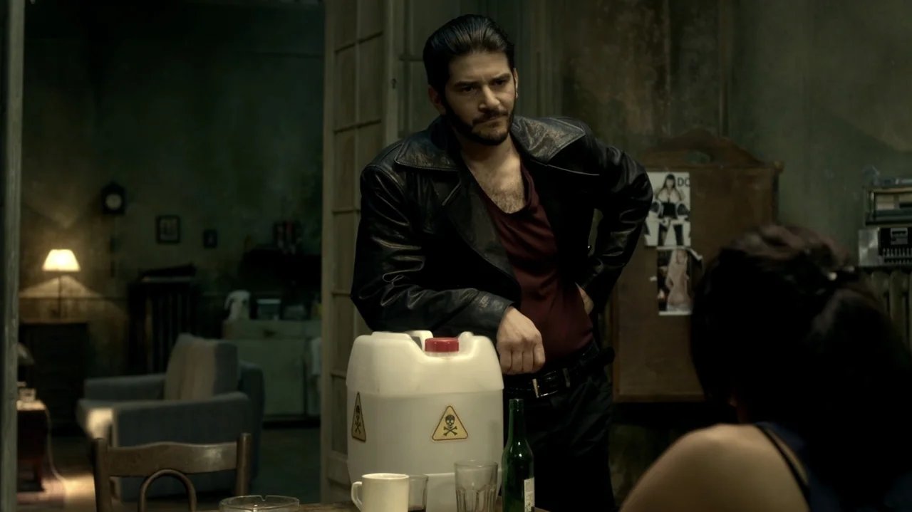 6 Bullets (2012) movie still: medium — A dimly lit room with peeling walls and clutter in the background. A man stands at a table next to a large…