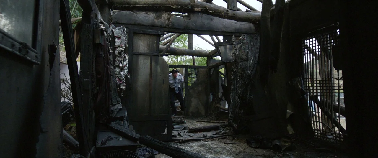 The Wailing (2016) movie still: wide — A fire-damaged wooden structure with exposed beams and charred debris scattered across the floor. A man in a…