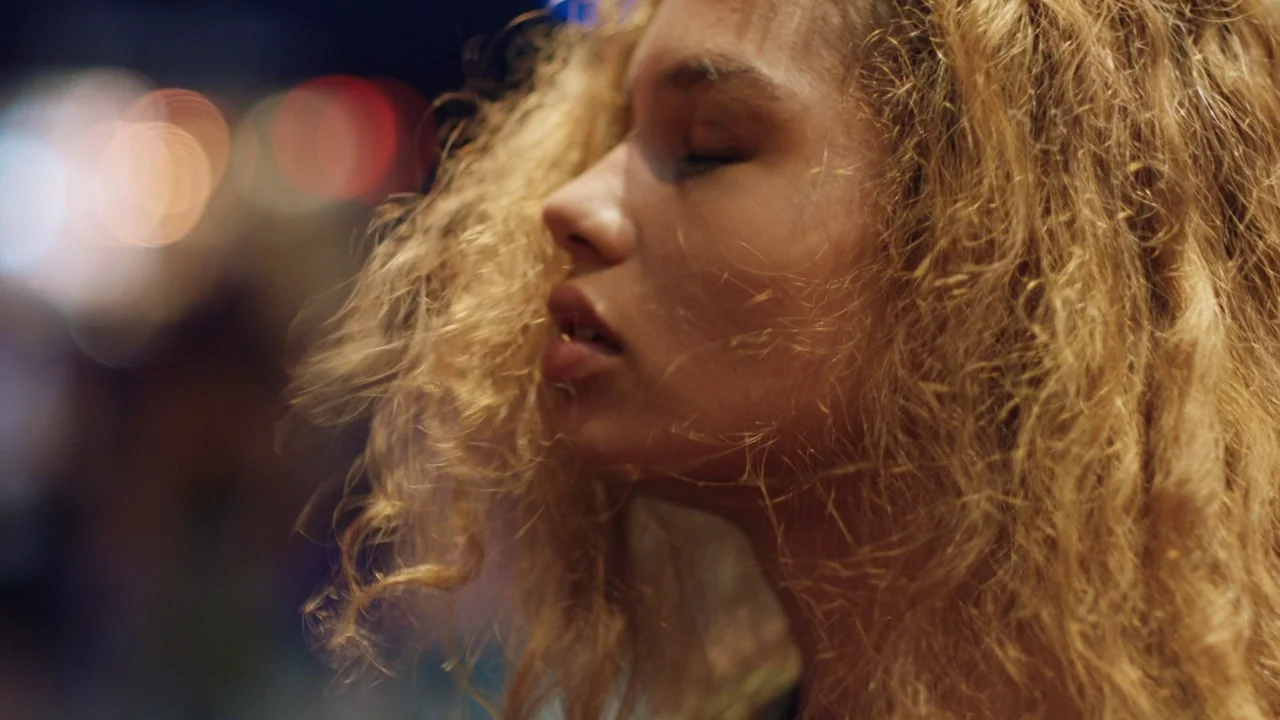 Madeline’s Madeline (2018) movie still: closeup — A woman stands on a city street at night with a blurred urban backdrop. Her eyes are closed while…