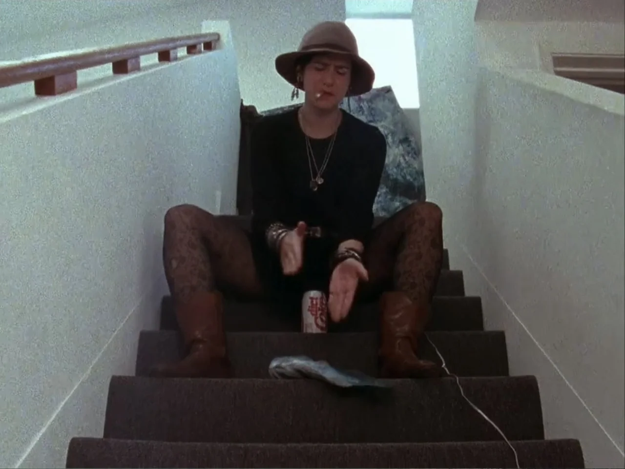 The Living End (1992) movie still: medium — A residential staircase with white walls and a dark carpeted floor. A woman in a hat sits on the steps…