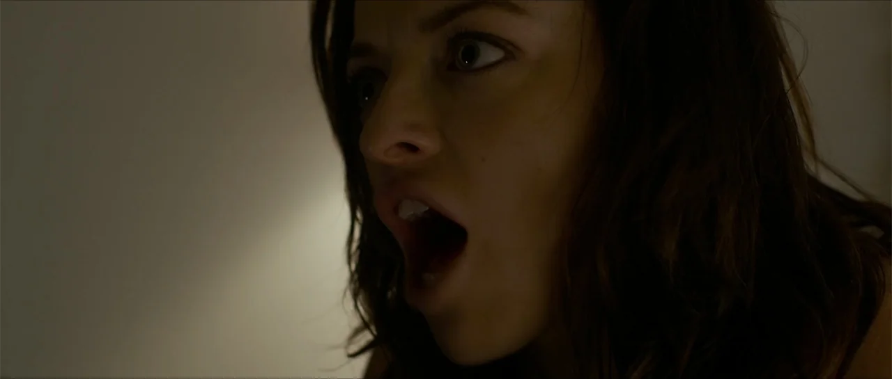 The Vault (2017) movie still: closeup — A dark indoor setting with a neutral wall background. A young woman looks off-screen with her mouth wide open.