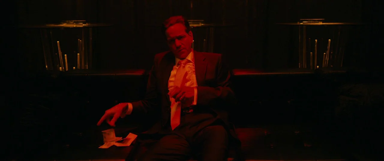 Hustlers (2019) movie still: medium — A man in a dark suit sits on a black leather couch in a lounge. He drops several banknotes from his hand onto…