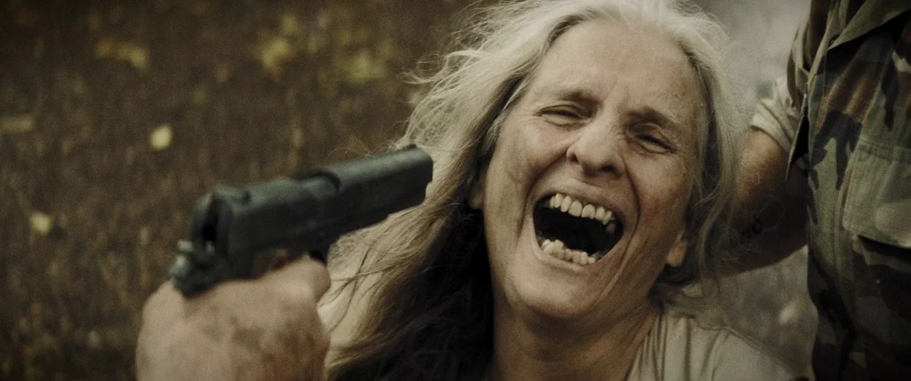 La Llorona (2019) movie still: closeup — A woman stands in a woodland area with a gun pointed directly at her. She is screaming with her mouth held…