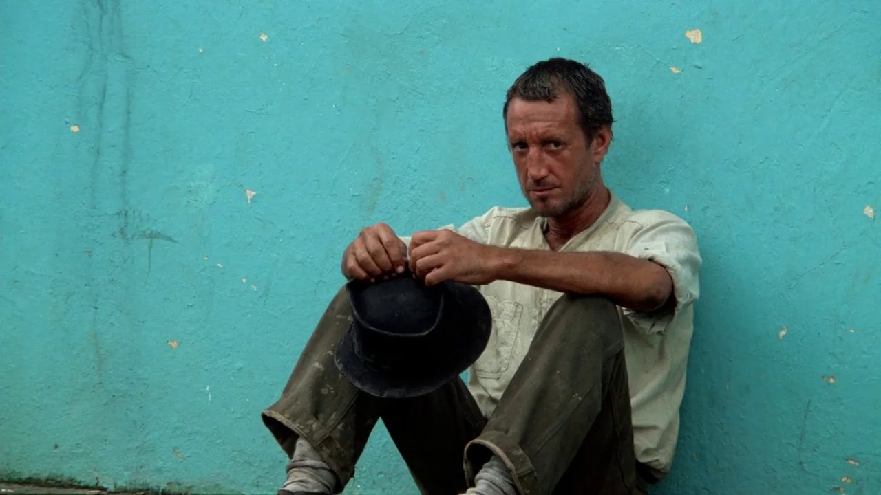 Sorcerer (1977) movie still: medium — A man sits on the ground in front of a textured, chipped turquoise wall. He holds a dark felt hat between his…