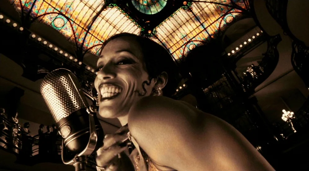 Frida (2002) movie still: closeup — A high-ceilinged hall features an expansive stained-glass skylight and dark, ornate architecture. A woman stands…