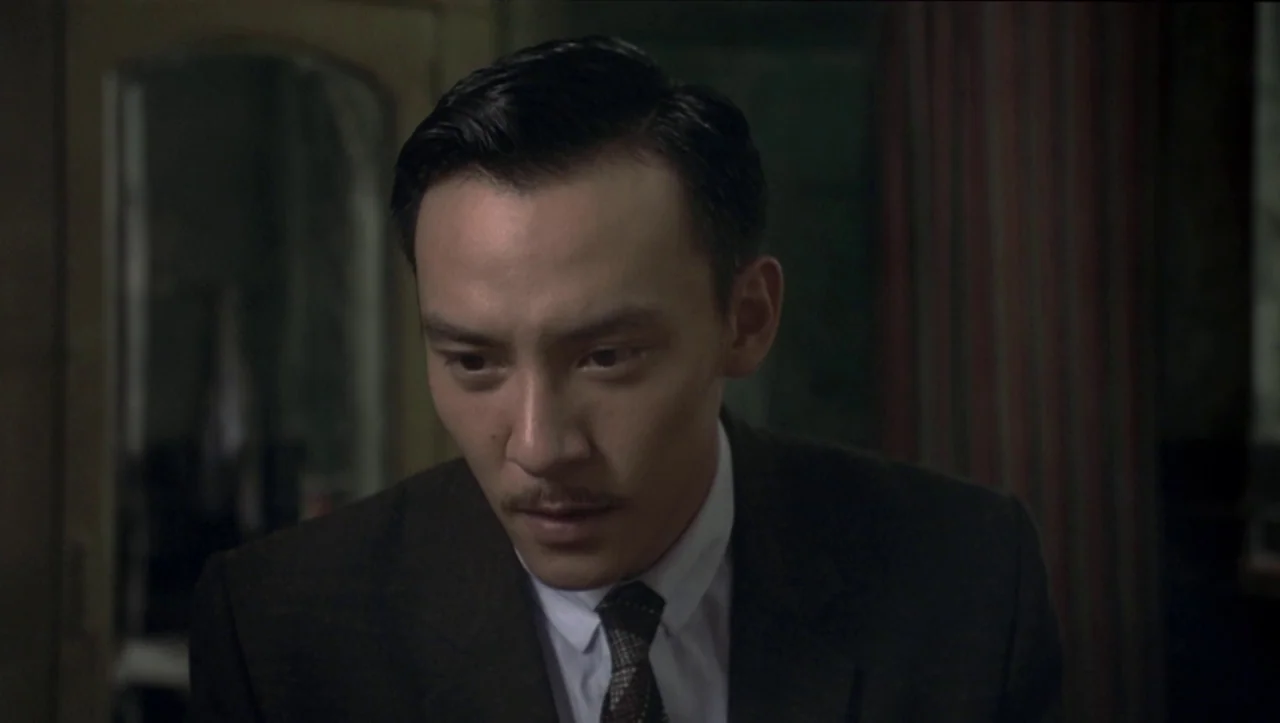 Eros (2004) movie still: closeup — A dimly lit room features dark wood paneling and indistinct vintage decor in the background. A man wearing a suit…