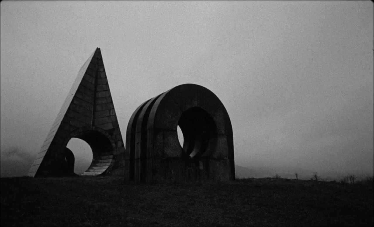 Last and First Men (2020) movie still: wide — A grassy hill holds two large geometric concrete structures under a featureless grey sky. The monument…