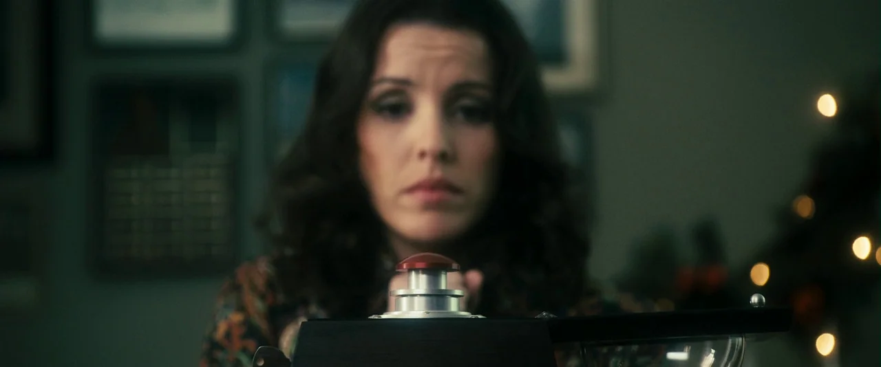 The Box (2009) movie still: medium — A dimly lit room with framed pictures on the wall and festive bokeh lights. A woman in a dark patterned blouse…