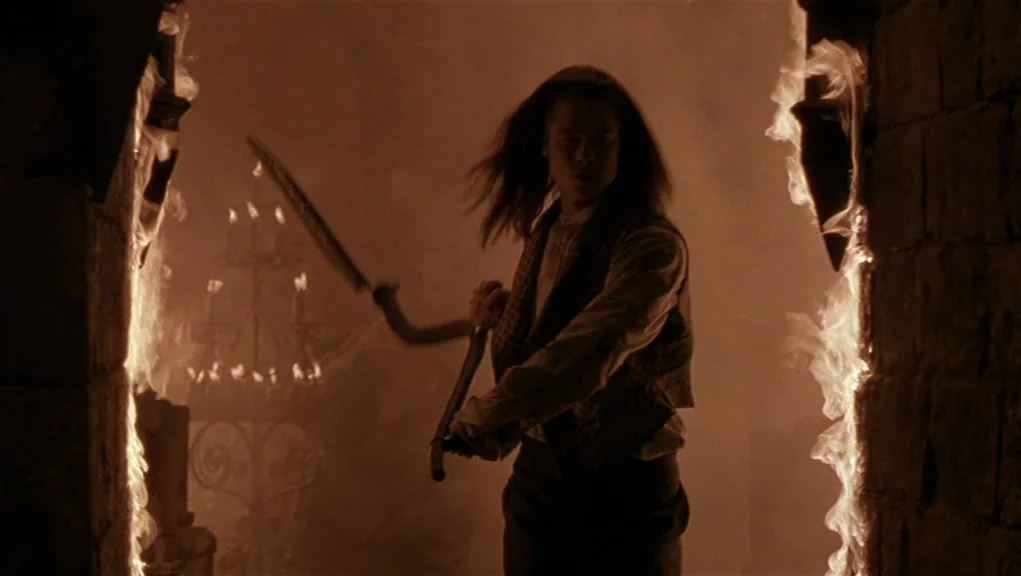 Interview With The Vampire movie still: medium — A dark stone passageway is framed by active flames climbing up the walls. A long-haired man holds a…