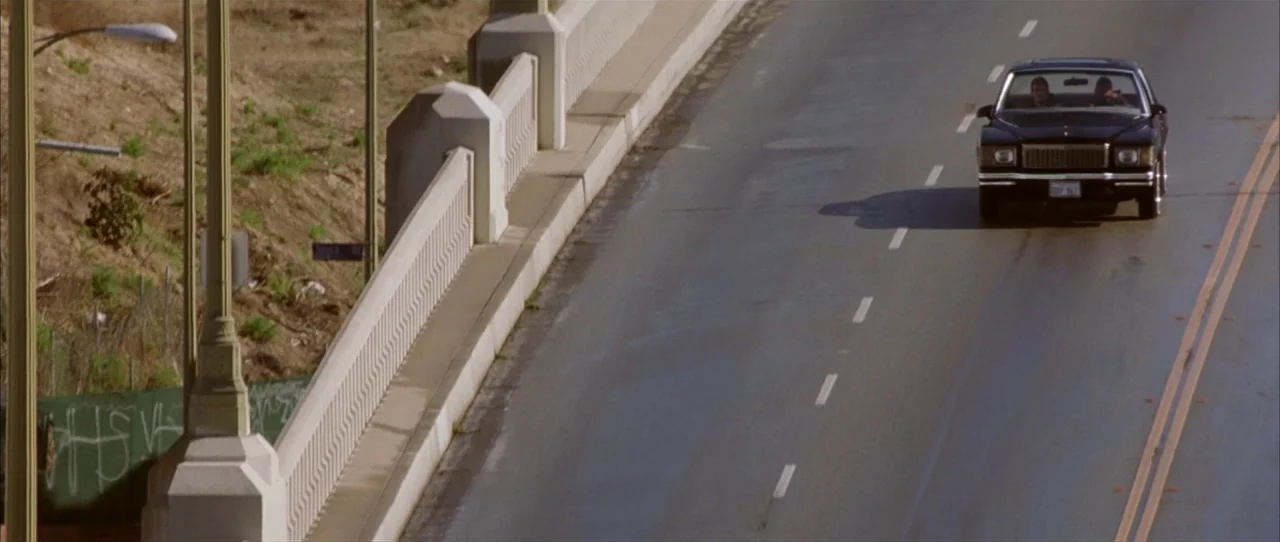 Training Day (2001) movie still: wide — A dark-colored sedan travels along a concrete bridge with a painted lane divider. The bridge railing runs…
