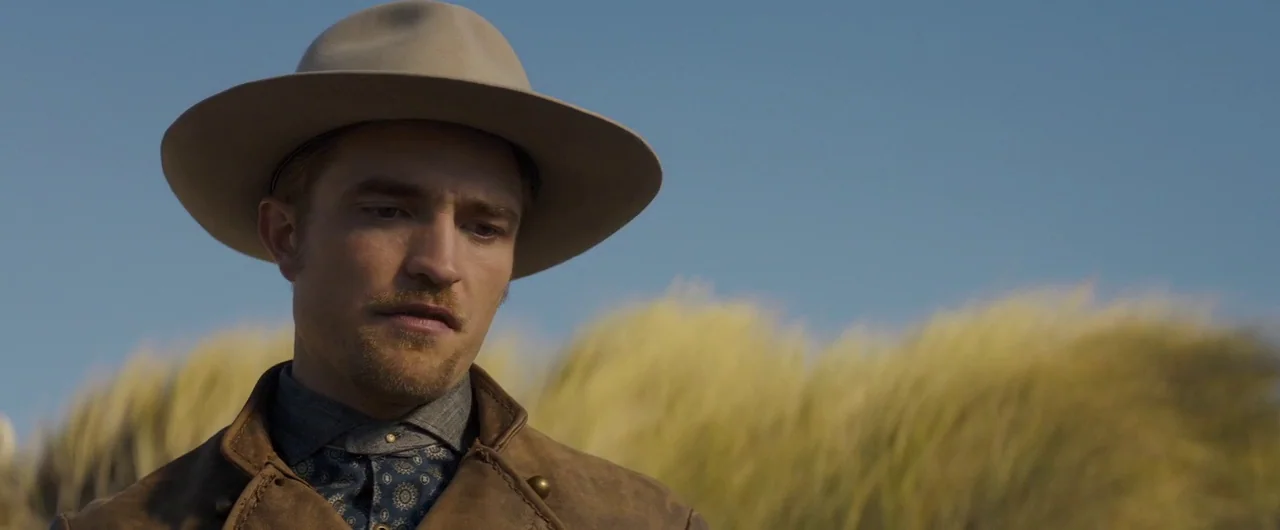 Damsel (2018) movie still: closeup — A coastal landscape with tall, sun-bleached dune grasses under a clear blue sky. A man wears a wide-brimmed…